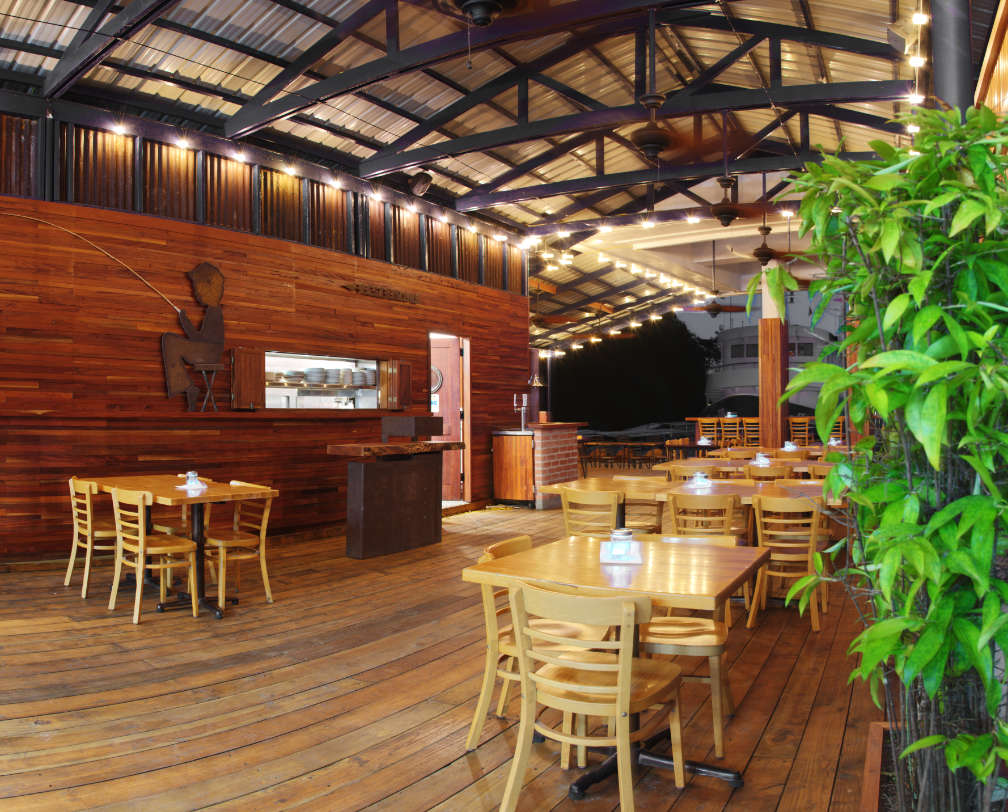 Inside Barracuda Restaurant, showing tables and chairs, looking towards the kitchen with a small window to deliver food and the sea at the right edge of the photo